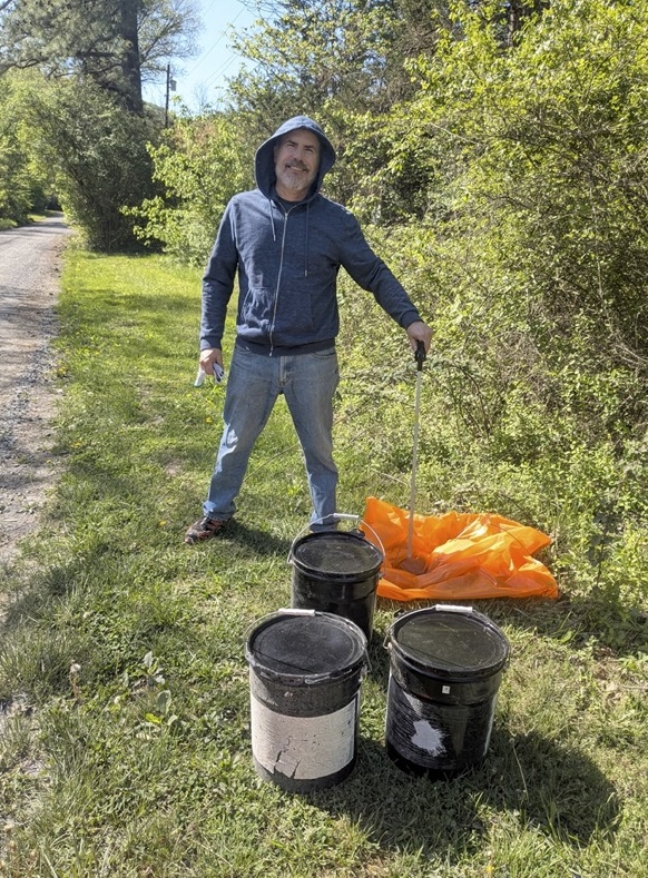A man in a blue hoodie and jeans stands on a grassy path beside three black barrels and an orange tarp.
