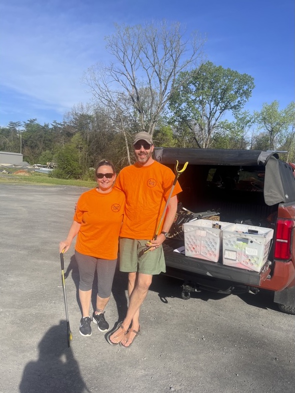 Two people in bright orange shirts stand beside a pickup truck with crates in its bed on a sunny day.